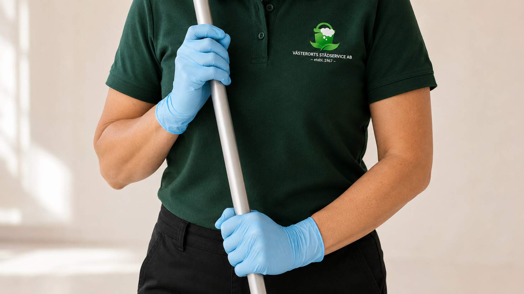 Städare i grön piké med Västerorts Städservice logotyp håller ett moppskaft i blå skyddshandskar